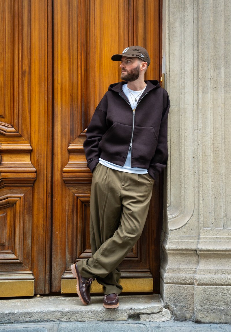 Sweat à capuche marron zippé par-dessus une chemise blanche, pantalons vert olive, chaussures marron à lacets. Debout contre une porte en bois avec des gravures détaillées.