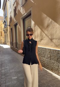 Młoda kobieta w czarnej bezrękawnikowej bluzce i beżowych spodniach, spacerująca w świetle dziennym po wąskiej kamienistej uliczce otoczonej beżowymi budynkami.