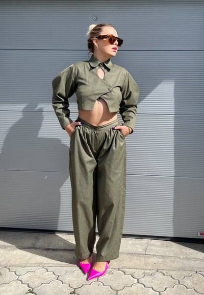 Chaqueta corta de color verde oliva con un detalle de agujero y pantalones de pierna ancha, combinados con tacones de punta rosa brillante. Textura de tela suave.