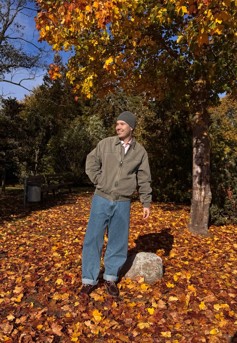 Olivengrøn bomberjakke, lyserød skjorte, løse blå jeans og mørkt fodtøj. Omgivet af efterårsblade og træer.