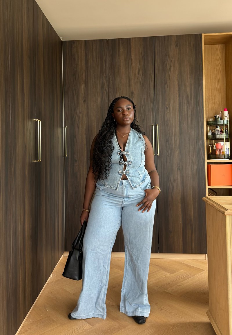Femme aux longs cheveux bouclés portant un gilet en denim bleu clair et un jean à jambes larges, tenant un sac noir, debout dans une pièce avec des armoires en bois.