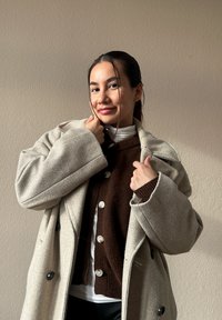 Ung kvinna med mörkt hår som bär en beige kappa och en brun knäppt tröja, ler mot en enfärgad ljusbrun vägg.