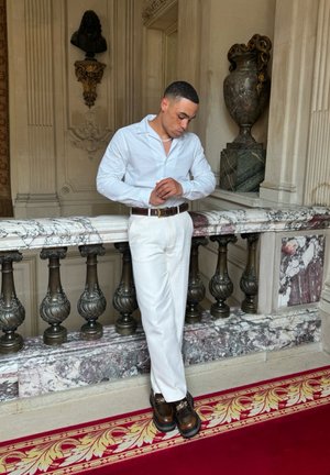 Camisa blanca texturizada, pantalones blancos, cinturón marrón y zapatos oscuros con acentos dorados. Barandilla de mármol y decoración ornamental en un interior clásico.