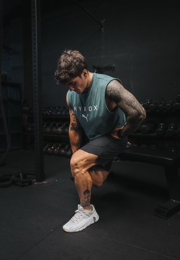 Homme avec des tatouages portant un débardeur et un short effectuant un exercice penché dans une salle de sport avec des poids en arrière-plan.