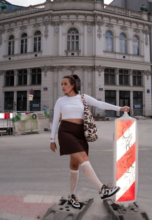 Brown skirt, white long-sleeve crop top, patterned shoulder bag. Wearing lace thigh-highs and black-and-white sneakers. Red and white barrier nearby.