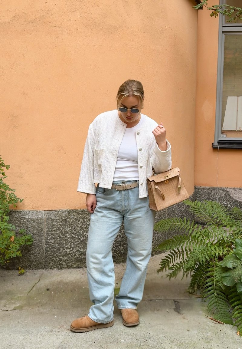 Hvid tekstureret jakke, hvid T-shirt, lyseblå baggy jeans, beige bælte, lysebrune slippers, holder en beige håndtaske med guldaccenter.