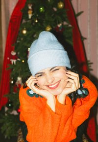 Personne souriante en pull orange et bonnet bleu clair reposant son menton sur ses mains, avec un sapin de Noël décoré en arrière-plan.