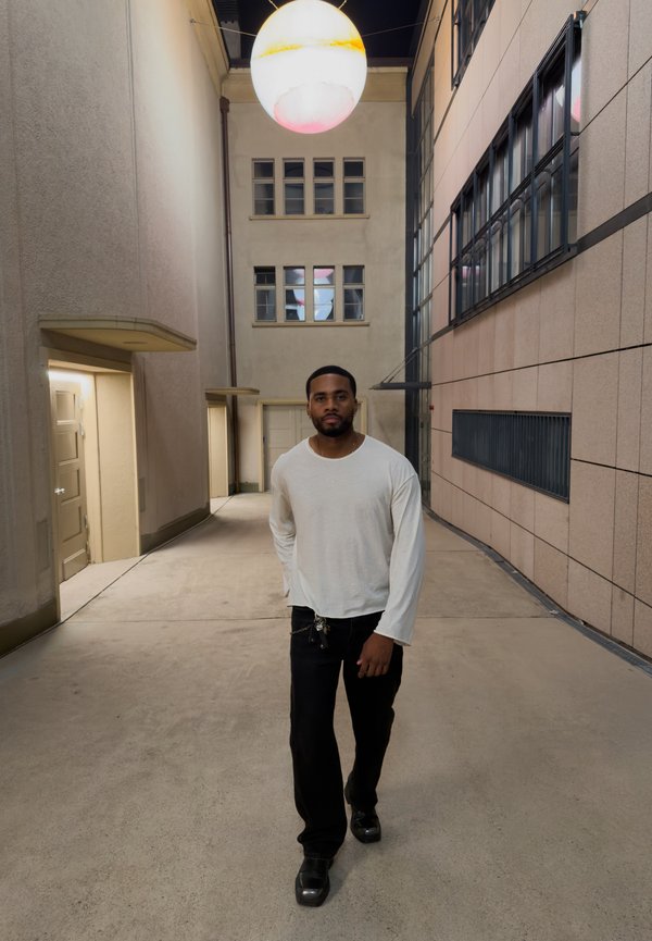 Un hombre con una camisa blanca y pantalones negros camina por un estrecho patio interior bajo una luminaria esférica iluminada.