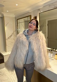 Mīksts, gaiši beidžs mākslīgais kažoks ar plašām piedurknēm, virs pelēka rāvējslēdzēja jaku, apvienots ar mirdzošiem šortiem un pelēkajām zeķbiksēm vannas istabā.