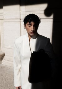 Hvit blazer med høyt halsdesign, båret over en svart håndveske. Scenen har sterke kontraster mellom lys og skygge med en teksturert steinbakgrunn.