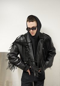 Veste de motard en cuir noir avec des franges sur les manches, fermeture éclair argentée, portée sur un sweat à capuche noir, associée à un pantalon foncé et des lunettes de soleil.