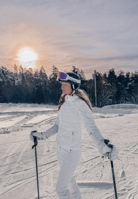 Femme en tenue de ski blanche debout sur une pente enneigée, tenant des bâtons, avec le soleil couchant derrière des arbres couverts de neige en arrière-plan.