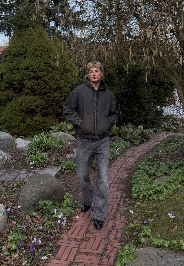 Ung mand med blondt hår og briller iført en mørk jakke og grå jeans, der går på en brostenssti i en have med grønne buske og små lilla blomster.