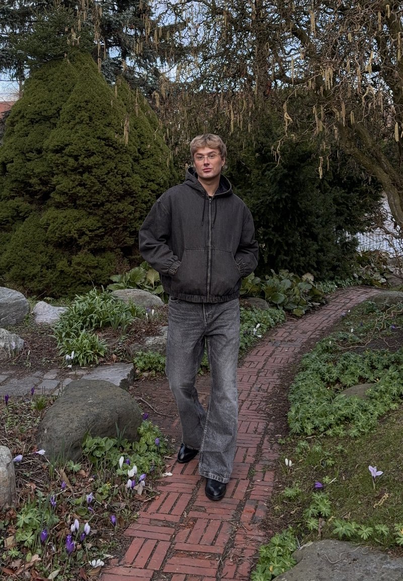 Jeune homme aux cheveux blonds et lunettes, portant une veste foncée et un jean gris, marchant sur un chemin en briques dans un jardin avec des buissons verts et de petites fleurs violettes.