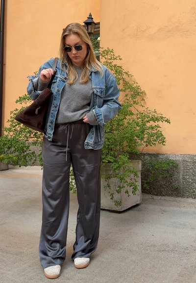 Chaqueta de mezclilla sobre jersey gris, pantalones satén grises y zapatos beige. Los accesorios incluyen un bolso de hombro marrón y gafas de sol negras. Texturas suaves visibles.