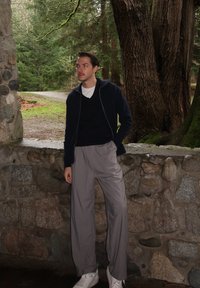 Pull à fermeture éclair bleu foncé, pantalon plissé gris et baskets blanches. Mur en pierre en arrière-plan avec une zone boisée derrière. Éclairage naturel.