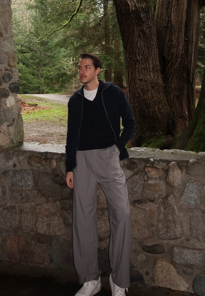 Suéter azul oscuro con cremallera, pantalones plisados grises y zapatillas blancas. Fondo de pared de piedra con un área boscosa detrás. Iluminación natural.