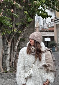 Biely nadýchaný kabát z ovčej vlny, béžová pletená čiapka s nápisom "WE", vzorovaný hnedý šál, malá biela crossbody taška, pozadie z tehlovej steny a zeleň.