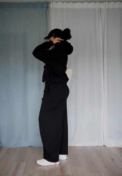 Sudadera negra cropped y pantalones anchos negros combinados con zapatillas blancas. La figura está posicionada de lado contra un fondo claro.