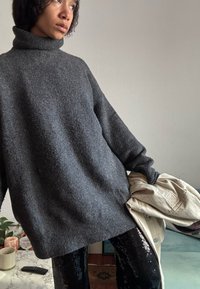Personne portant un pull à col roulé gris foncé oversize et un pantalon noir à paillettes, tenant une veste beige clair dans une pièce minimalement décorée.