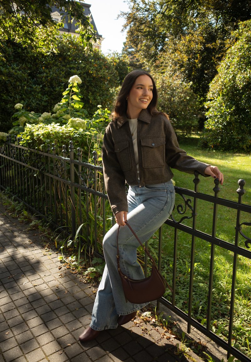 Jaqueta castanha curta com dois bolsos no peito, t-shirt cinza, jeans azul claro com boca de sino, bolsa castanha e botas de tornozelo, ambientada num parque verde.