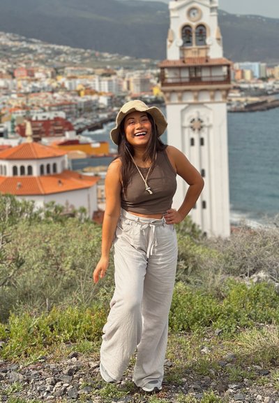 Joven sonriente posando al aire libre cerca de vegetación con ciudad costera y torre del reloj al fondo.