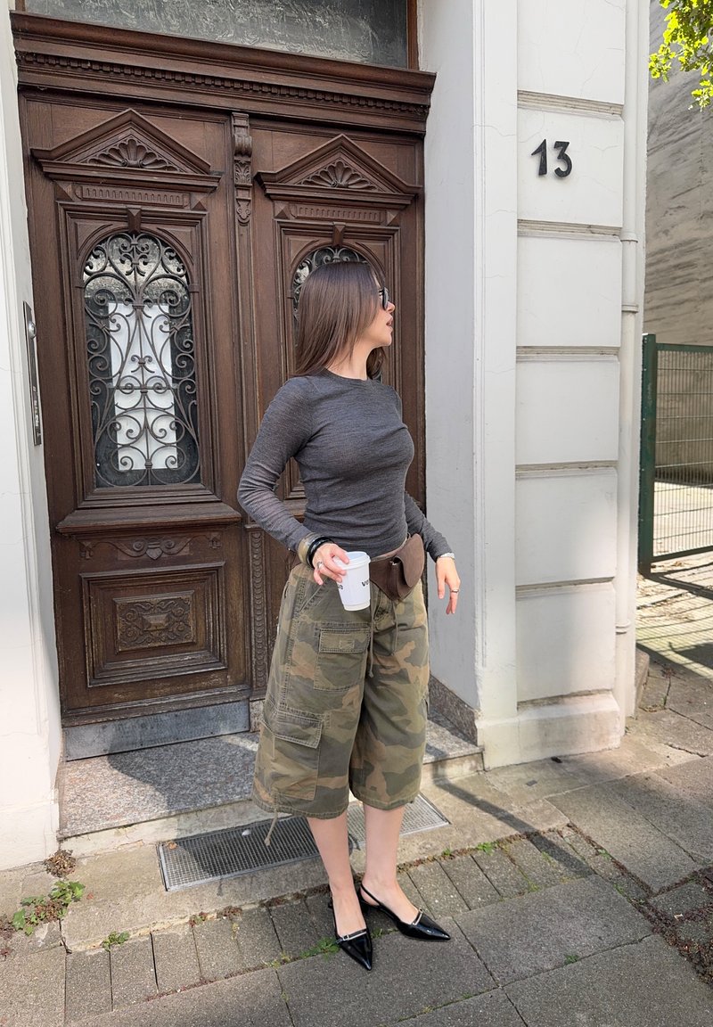 Femminile con capelli lunghi, occhiali da sole, maglietta grigia a maniche lunghe, pantaloni cargo mimetici e scarpe nere a punta, mentre tiene una tazza di caffè vicino a una porta di legno.
