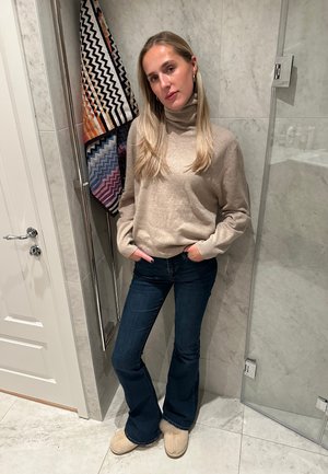 Jersey beige con cuello alto, jeans acampanados de color azul oscuro y zapatillas de color marrón claro. El fondo presenta toallas con patrones geométricos y una ducha de cristal.