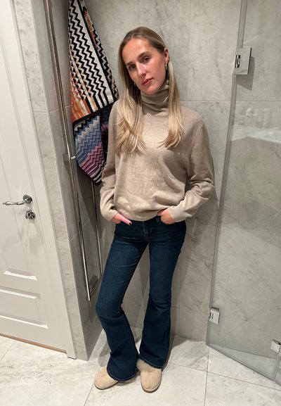 Jersey beige con cuello alto, jeans acampanados de color azul oscuro y zapatillas de color marrón claro. El fondo presenta toallas con patrones geométricos y una ducha de cristal.