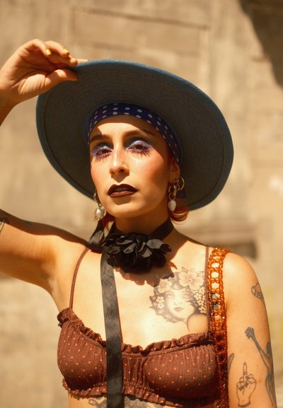Top corto de lunares marrón, collar de cinta negra, sombrero de ala ancha azul, bolso de crochet naranja, tatuajes coloridos y maquillaje atrevido con sombra de ojos morada.