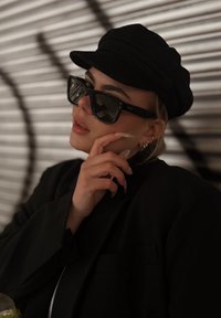 Fekete napszemüveg téglalap alakú kerettel, fekete, szalaggal díszített, testhezálló sapka, valamint hosszú, hegyes körmök. A dzseki is fekete.