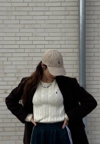 Femme portant une casquette beige, un pull en maille torsadée blanc, un blazer noir et un pantalon foncé, debout contre un mur en briques claires, les mains sur les hanches.