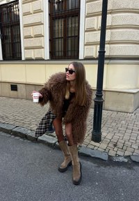 Femme en manteau en fourrure marron, jupe à carreaux et lunettes de soleil tenant une tasse, se penchant sur un trottoir pavé près d'un lampadaire.