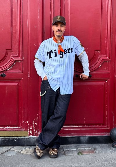 Una camiseta de béisbol azul claro con la inscripción "Tigers", pantalones de mezclilla oscuros, zapatos de estampado de leopardo y un pañuelo naranja contra una pared roja.