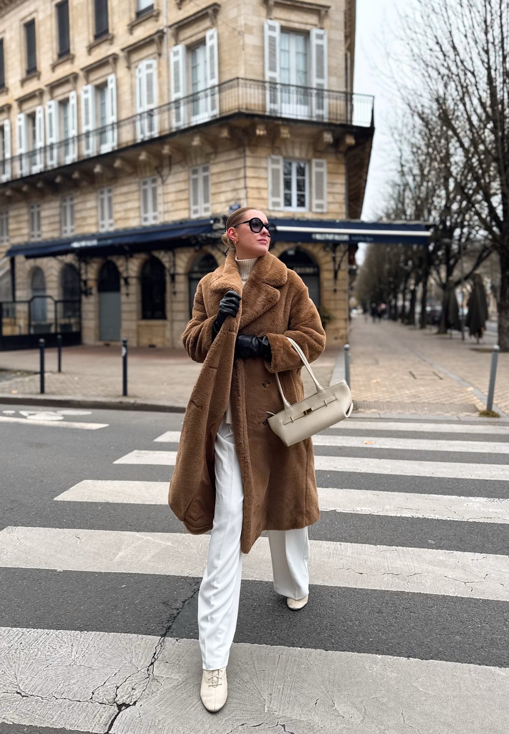 Manteau en fausse fourrure marron, col roulé blanc, pantalon blanc large et sac beige clair. Le modèle porte des lunettes de soleil foncées et des gants noirs sur un passage pour piétons.