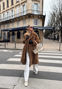 Manteau en fausse fourrure marron, col roulé blanc, pantalon blanc large et sac beige clair. Le modèle porte des lunettes de soleil foncées et des gants noirs sur un passage pour piétons.