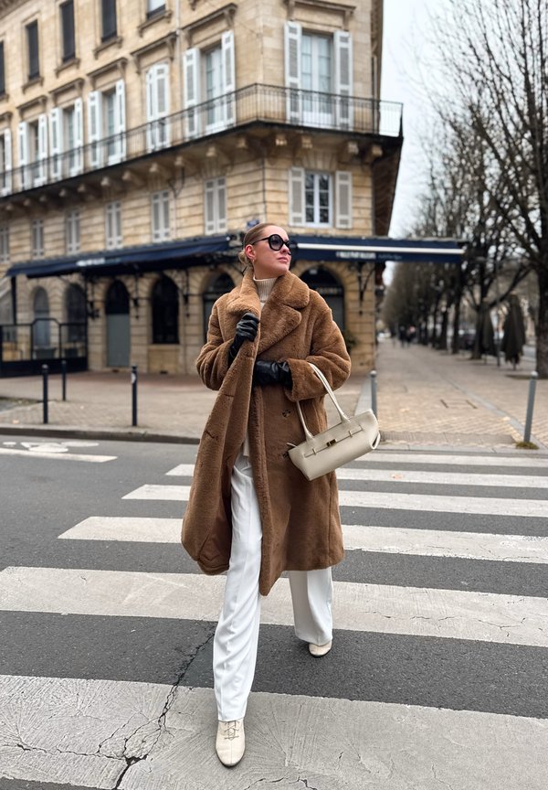 Manteau en fausse fourrure marron, col roulé blanc, pantalon blanc large et sac beige clair. Le modèle porte des lunettes de soleil foncées et des gants noirs sur un passage pour piétons.