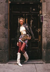 Schwarze Bomberjacke mit Logo, weißes eng anliegendes Top, gemusterter Minirock, rote Spitzenstrumpfhose und weiße strukturierte kniehohe Stiefel, die eine Tasche mit Schlangenprint hält.