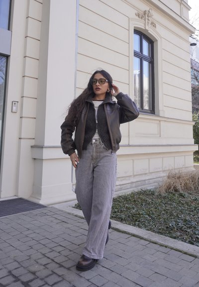 Mujer con chaqueta de cuero oscuro y vaqueros anchos grises caminando por un camino pavimentado junto a un edificio beige con una ventana grande.