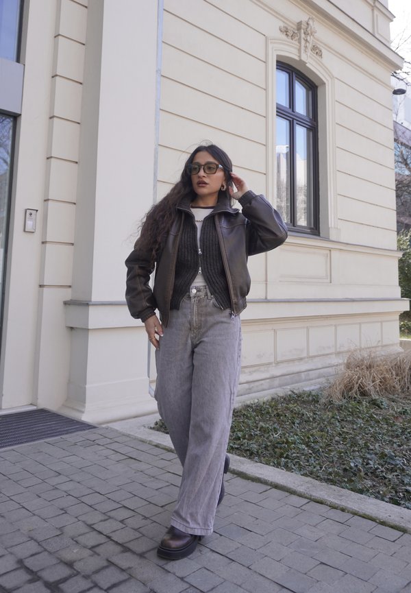 Mujer con chaqueta de cuero oscuro y vaqueros anchos grises caminando por un camino pavimentado junto a un edificio beige con una ventana grande.