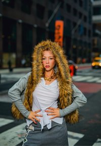 Naine, kes kannab karvase kapuutsiga vestit, valget lahtist sarnast pluusi ja halli pükse, seisab enesekindlalt käed puusas linnatänaval.