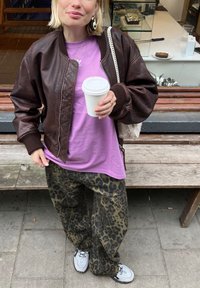 Jachetă bomber din piele maro, maiou violet, pantaloni cu imprimeu de leopard, adidași gri. Ținând o cană albă de cafea, cu o geantă texturată.