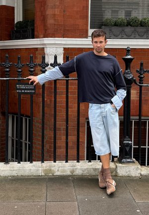 Blusa suelta negra y azul con mangas largas, pantalones cortos de mezclilla clara y zapatos marrones con cordones, frente a una pared de ladrillos con barandillas.