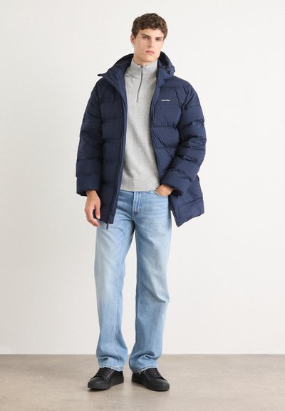 Chaqueta de plumas color marino con capucha, diseño acolchado, cierre con cremallera, llevada sobre un suéter de cuello alto gris claro, combinada con jeans azul claro y zapatillas negras.