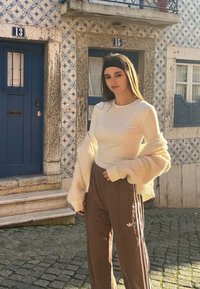Modelka má na sebe priliehavé biele tričko s dlhým rukávom a hnedé športové nohavice s bielymi pruhmi. Pozadie tvorí retro modré dvere a vzorované dlaždice.