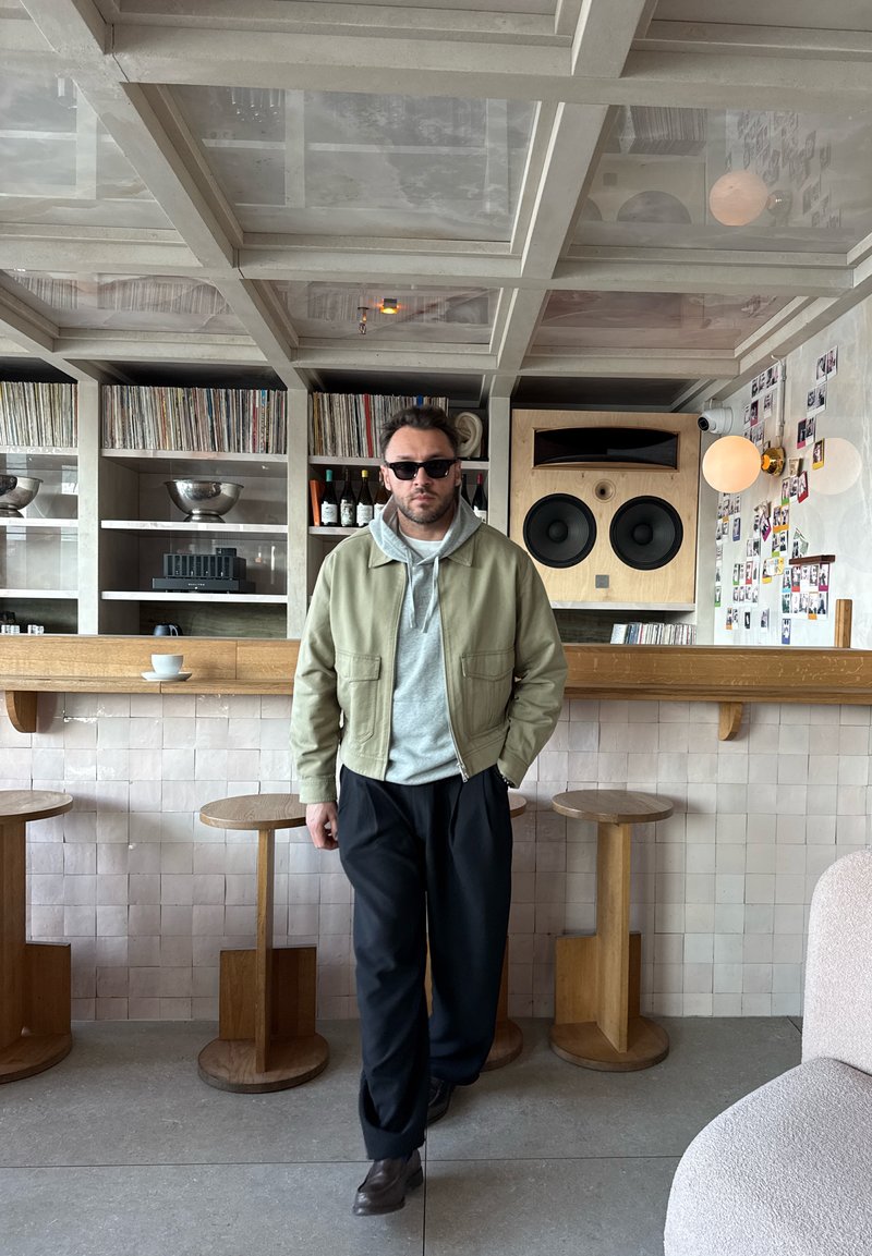 Homme portant des lunettes de soleil, une veste beige, un sweat à capuche gris et un pantalon noir marchant dans un café moderne avec des tabourets en bois et un grand haut-parleur sur le mur.