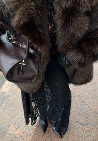 Brun pelsfrakk, svart blonderkjole, spisse svarte hæler og en mørk brun skinnveske med gullbeslag. Strukturert og lagdelt utseende.