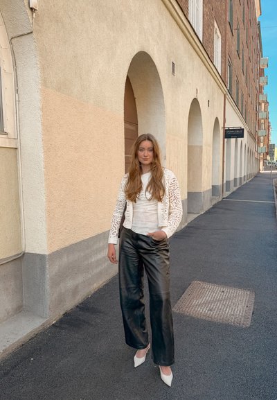 Joven mujer con el pelo largo que lleva una chaqueta blanca de encaje, top blanco, pantalones de cuero negros y tacones blancos, de pie en la acera urbana junto a unos arcos de edificio beige.