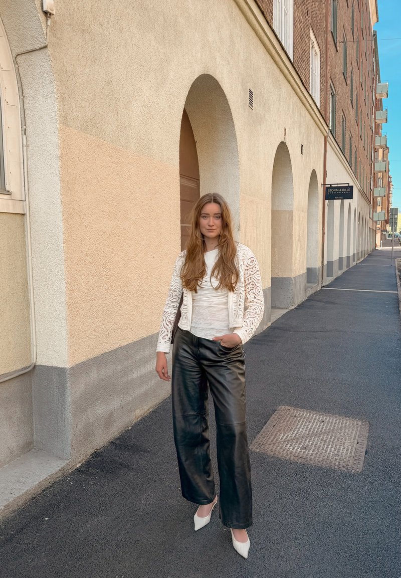 Jeune femme aux longs cheveux portant une veste en dentelle blanche, un top blanc, un pantalon en cuir noir et des talons blancs, debout sur un trottoir urbain près des arches d’un bâtiment beige.