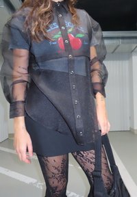 Crna prozirna košulja s dugim rukavima i grafičkim uzorkom, složena preko crne haljine, u paru s mrežastim čarapama. Suptilni sjaj tkanine.